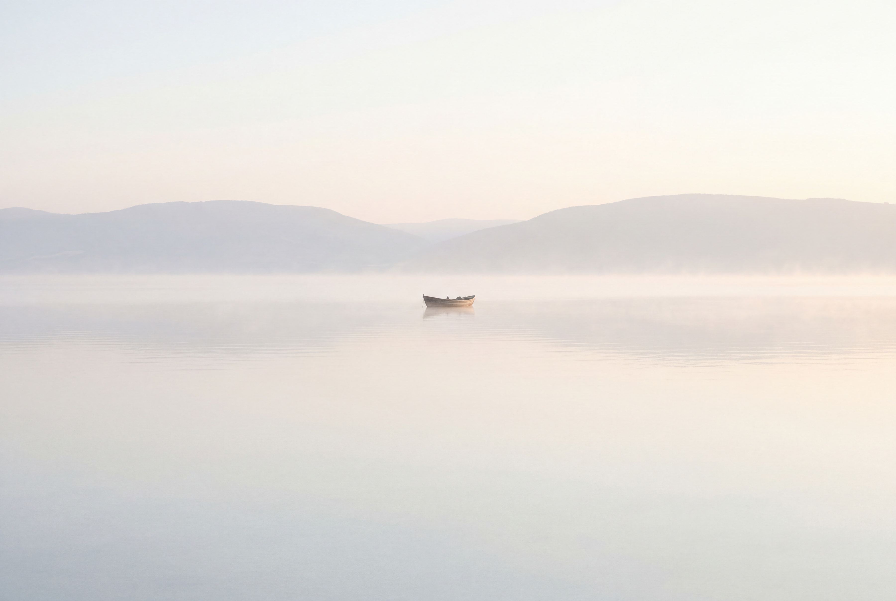 The Sea of Galilee at first light, glass-still under a pale mist, with one small wooden boat far out on the water.