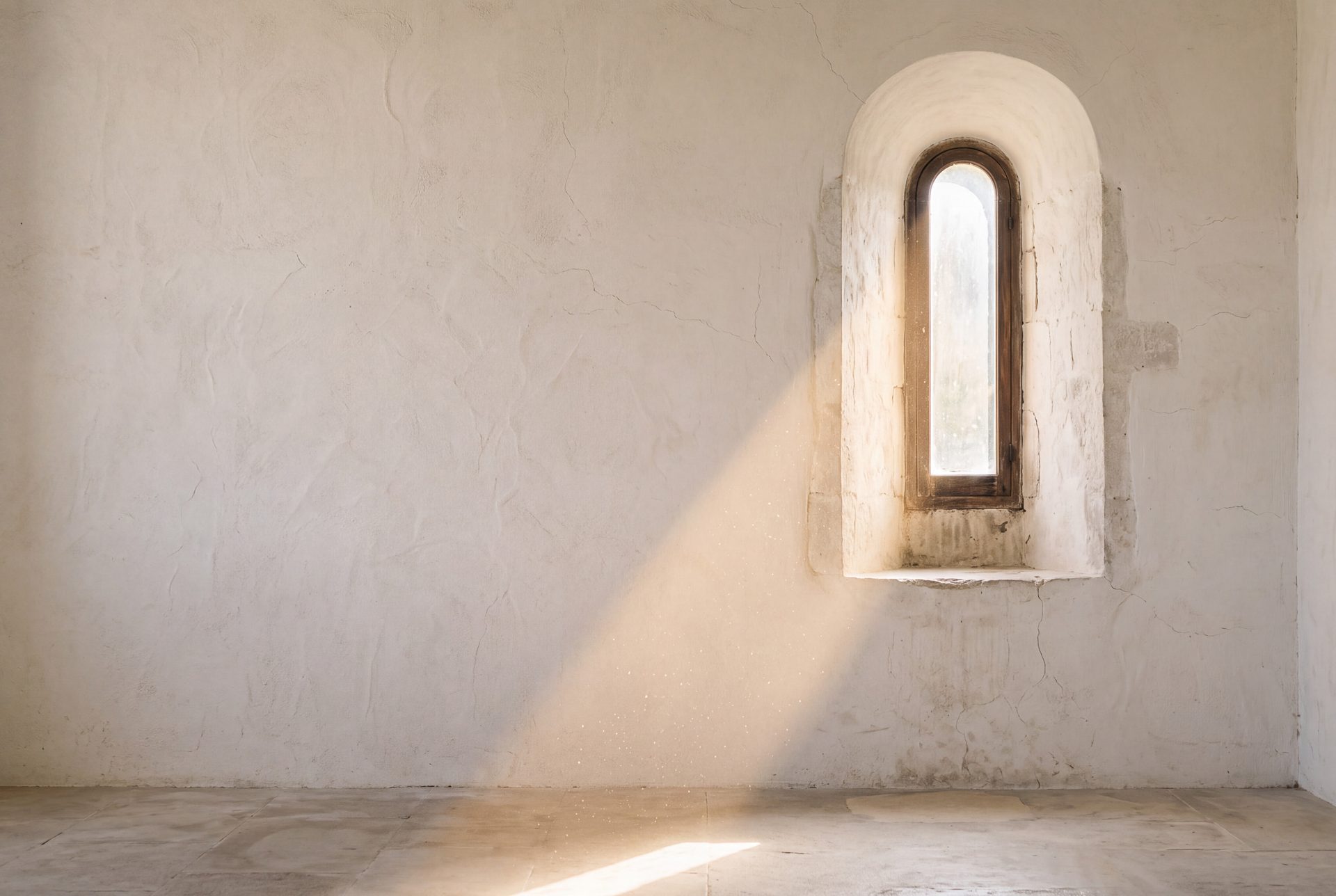Morning sun falling through a narrow arched window across a limewashed stone wall and floor.